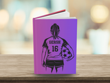 A hardcover notebook or journal standing upright on a wooden surface, featuring a purple-to-pink gradient cover with a black graphic of a soccer player from the back holding a ball. The cover has the text 'GERSH 16' printed on it.