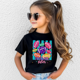 Little girl standing in bleachers holding a football. She's wearing a black t-shirt with a colorful design: teal vertical stripes, pink, purple, yellow and orange footballs scattered around. Inside the balls are lighting bolts, flowers, hearts, leopard print, etc. At the bottom in pink and purple it says Football Sister.