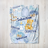 A personalized baby blanket with a light blue and white pattern featuring the name 'Sebastian' and illustrations of a teddy bear, baby gear, and toys, laid flat on a light wood background.