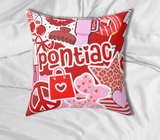 A square decorative throw pillow with a red, pink, and white graphic collage featuring the camp 'pontiac' in large white letters, hearts, stars, and candy illustrations, resting on white bedding.