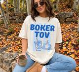 An off-white short-sleeve t-shirt featuring the blue text BOKER TOV above a graphic of an iced coffee and a hot tea cup. The design includes vertical light blue stripes behind the text and graphics. The shirt is worn with blue jeans.