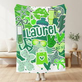 A throw blanket in shades of green featuring pop culture graphics like lip prints, sunglasses, peace signs, and the camp name 'Laurel'.