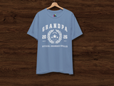A light blue short-sleeve t-shirt hanging on a wooden hanger against a dark wood plank background. The shirt features a white graphic print on the front with the text 'GRANDPA 20 26 OFFICIAL GRANDKID SPOILER' surrounding a star and wreath design.