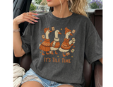 A woman wearing a dark gray graphic t-shirt with a cartoon design of three geese in orange sweaters and the text 'IT'S TILE TIME'.