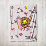 A personalized softball-themed blanket featuring the name 'COLLEEN' repeated, decorated with illustrations of pink bows, pink hearts, yellow softballs, a brown softball glove, and a pink drink cup, displayed on a light wood plank background.