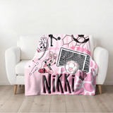 A pink and white soccer-themed personalized throw blanket draped over a white armchair in a bright room. The blanket features the name 'Nikki' in large black letters, a soccer ball, a soccer goal, a soccer glove, cleats, a pink and white patterned background.