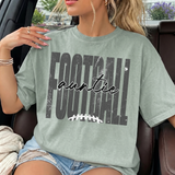 A woman wearing a light sage green graphic t-shirt with a distressed 'FOOTBALL' text design and the word 'auntie' in script, sitting in a car.