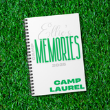A spiral-bound notebook titled "Ellie's Memories 2026" with "Camp Laurel" printed on the cover, placed on bright green artificial grass.