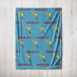 A personalized blue blanket with a repeating pattern of red 'ASHLEY' text, yellow softballs, and wooden bats, laid flat on a light wood plank background.