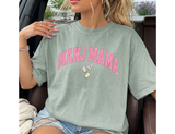 Woman sitting wearing a light green Comfort Colors t-shirt that says 'MAHJ MAMA' in a pink, distressed, varsity arched font. 3 Mahjong tiles underneath. 
