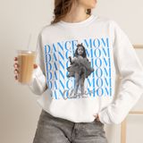 A woman wearing a white crewneck sweatshirt with a blue 'Dance Mom' graphic and a black-and-white photo of a young girl, holding an iced coffee in a light neutral indoor setting.