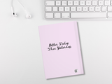 A light pink notebook with the black cursive text 'Better Today Than Yesterday.' on the cover, styled on a white desk next to a white keyboard and white earbuds.