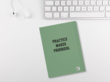 A sage green hardcover notebook with the text 'PRACTICE MAKES PROGRESS.' in black, styled on a white desk next to a keyboard and earbuds.