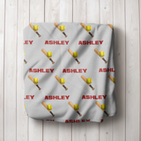 A personalized grey blanket with a repeating pattern of red 'ASHLEY' text, yellow softballs, and wooden bats, folded up. 