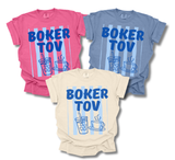 Three graphic t-shirts in pink, blue, and cream, all featuring a blue 'BOKER TOV' text design with striped background and a drink and coffee cup illustration.