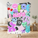 A colorful throw blanket featuring pop culture graphics like lip prints, sunglasses, peace signs, and the name 'Alana' in pink, held up by a person standing in a living room.