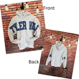 A light gray hoodie shown from both front and back on a hanger against a brick wall backdrop. The front has large navy blue 'TYLER HILL' lettering across the chest, and the back has two blue star graphics on the sleeves.