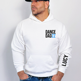 A man wearing a white pullover hoodie with 'DANCE DADZZ' printed in black and blue on the chest, and 'LUCY' printed in black on the left sleeve. He is wearing blue jeans and a black baseball cap, standing against a light gray wall.