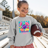 Little girl standing in bleachers holding a football. She's wearing a grey crewneck sweatshirt with a colorful design: teal vertical stripes, pink, purple, yellow and orange footballs scattered around. Inside the balls are lighting bolts, flowers, hearts, leopard print, etc. At the bottom in pink and purple it says Football Sister.