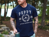 A man wearing a navy blue graphic t-shirt with a white 'Poppy 2026 Official Grandkid Spoil' design, standing outdoors in a wooded area and holding a dark green tumbler.