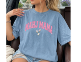 Woman sitting wearing a light blue Comfort Colors t-shirt that says 'MAHJ MAMA' in a pink, distressed, varsity arched font. 3 Mahjong tiles underneath. 