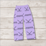 Light purple pants with an all-over repeating pattern of black crossed field hockey sticks and the name 'JORDYN', shown laid flat on a light wood plank background.