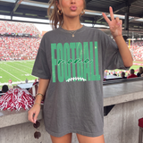 A woman in a stadium wearing an oversized gray t-shirt with 'FOOTBALL mama' printed in green and white, making a peace sign. The background shows a crowded football field and stadium seating.