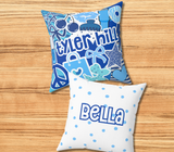 A square decorative throw pillow with blues and white graphic collage featuring the camp 'tyler hill' in large blue letters, hearts, stars, and candy illustrations. The back of the pillow is white with green polka dots and the name 'Bella' across the center in blue.