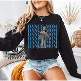 A woman wearing a black crewneck sweatshirt with a blue and white graphic print that reads 'Dance Mom' alongside a grayscale photo of a ballerina, paired with denim shorts in a cozy indoor setting.
