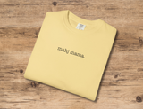 A folded pale yellow crewneck t-shirt with the text 'mahj mama.' printed in black serif font on the chest, displayed on a rustic wooden surface.