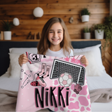 A smiling young girl with long light brown hair sits in a bedroom holding up a personalized pink soccer throw blanket. The blanket features a pink and white cow print pattern, a soccer ball graphic, a soccer goal, pink cleats, and the name 'Nikki' in large black letters.