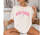 Woman sitting wearing an ivory Comfort Colors t-shirt that says 'MAHJ MAMA' in a pink, distressed, varsity arched font. 3 Mahjong tiles underneath. 
