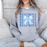 A woman wearing a heather gray hoodie with a blue graphic print that reads 'WESTMONT' repeated with a light blue bow in the center, sitting on a couch.