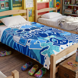 A throw blanket in shades of blue featuring pop culture graphics like lip prints, sunglasses, peace signs, and the camp name 'Tyler Hill' laid out on a camp bed. 