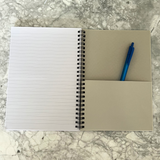 An open spiral-bound notebook with lined pages on the left and featuring a blue pen in a pocket on the right. on a gray marble surface.