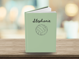 A personalized hardcover notebook with a sage green cover, featuring the name 'Stephanie' and a sketch of a volleyball in black ink, displayed on a wooden surface with a blurred indoor background.