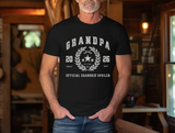 A man wearing a black graphic t-shirt with a white 'Grandpa Official Grandkid Spoiler' design, standing in a rustic wooden room.