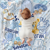 A baby wearing a white bodysuit lies on their belly on a light blue and white personalized baby blanket featuring illustrations of toys, stars, pacifiers, and the name 'Sebastian'.