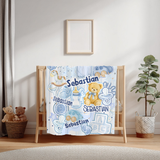 A personalized baby blanket with a blue and white watercolor design featuring the name 'Sebastian', a teddy bear, pacifiers, and baby accessories, draped over a wooden crib in a styled nursery setting.