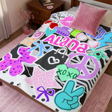 A throw blanket in vibrant colors like purple, pink and teal featuring pop culture graphics like lip prints, sunglasses, peace signs, and the name 'Alana' laid out on a bed. 