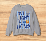 A heather gray crewneck sweatshirt featuring a festive Hanukkah graphic with the text 'LOVE LIGHT & LATKES' in dark blue lettering over light blue vertical stripes, accompanied by illustrations of a menorah, a dreidel, and a latkes displayed flat on a light wood background.