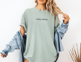 A woman wearing an oversized sage green t-shirt with 'mahj mama.' printed in black across the chest, styled with a light-wash denim jacket draped off her shoulders against a white background.