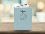 A personalized hardcover journal standing upright on a wooden surface. The cover is a light pastel blue with the name 'Alyssa' written in black script above a simple line drawing of a volleyball.