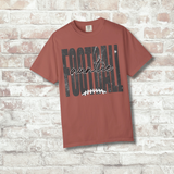A terracotta-colored short-sleeve t-shirt with a distressed black and white graphic reading 'FOOTBALL auntie' with a football laces detail, displayed against a white brick wall background.
