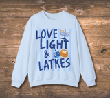 A light blue crewneck sweatshirt with a festive Hanukkah graphic reading 'LOVE LIGHT & LATKES' in dark blue lettering over light blue stripes, featuring illustrations of a menorah, dreidel, and latkes, displayed flat on a rustic wooden background.