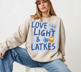 A woman wearing a beige crewneck sweatshirt with a blue and yellow 'Love Light & Latkes' graphic, paired with blue jeans, sitting against a plain white background.