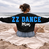 A woman sits on a beach blanket on the sand with her back to the camera, wearing a black sweatshirt with 'ZZ DANCE Mom' printed on the back in blue and white lettering.