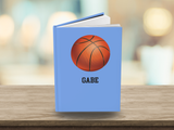 A personalized hardcover notebook with a light blue cover, featuring a printed orange basketball graphic and the name 'GABE' in black text, displayed on a wooden surface with a blurred background.