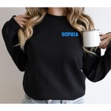 A woman wearing a black crewneck sweatshirt with the name 'SOPHIA' printed in bright blue on the chest, holding a white enamel mug.