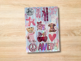 A personalized sticker sheet with a light blue and pink striped background, decorated with ballet-themed graphics, a teddy bear in pink, a heart, a peace sign, and the name 'Avery' in glittery letters, displayed on a light wood surface.